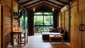 Interior de una cabaña de troncos con piso de madera, un sofá, una mesa, un televisor y un ventanal con vistas a árboles.