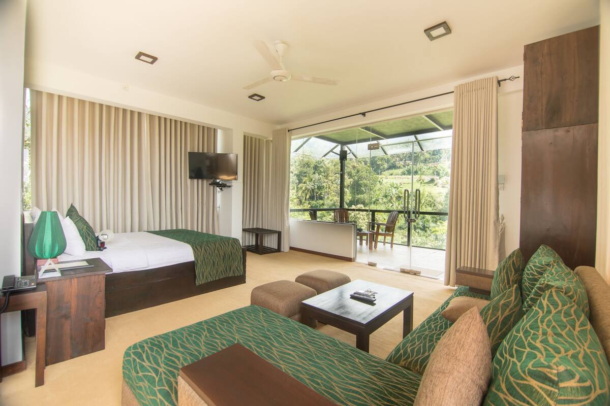 Quarto de hotel com cama, sofá, mesa de centro, televisão e varanda com vista para o verde.