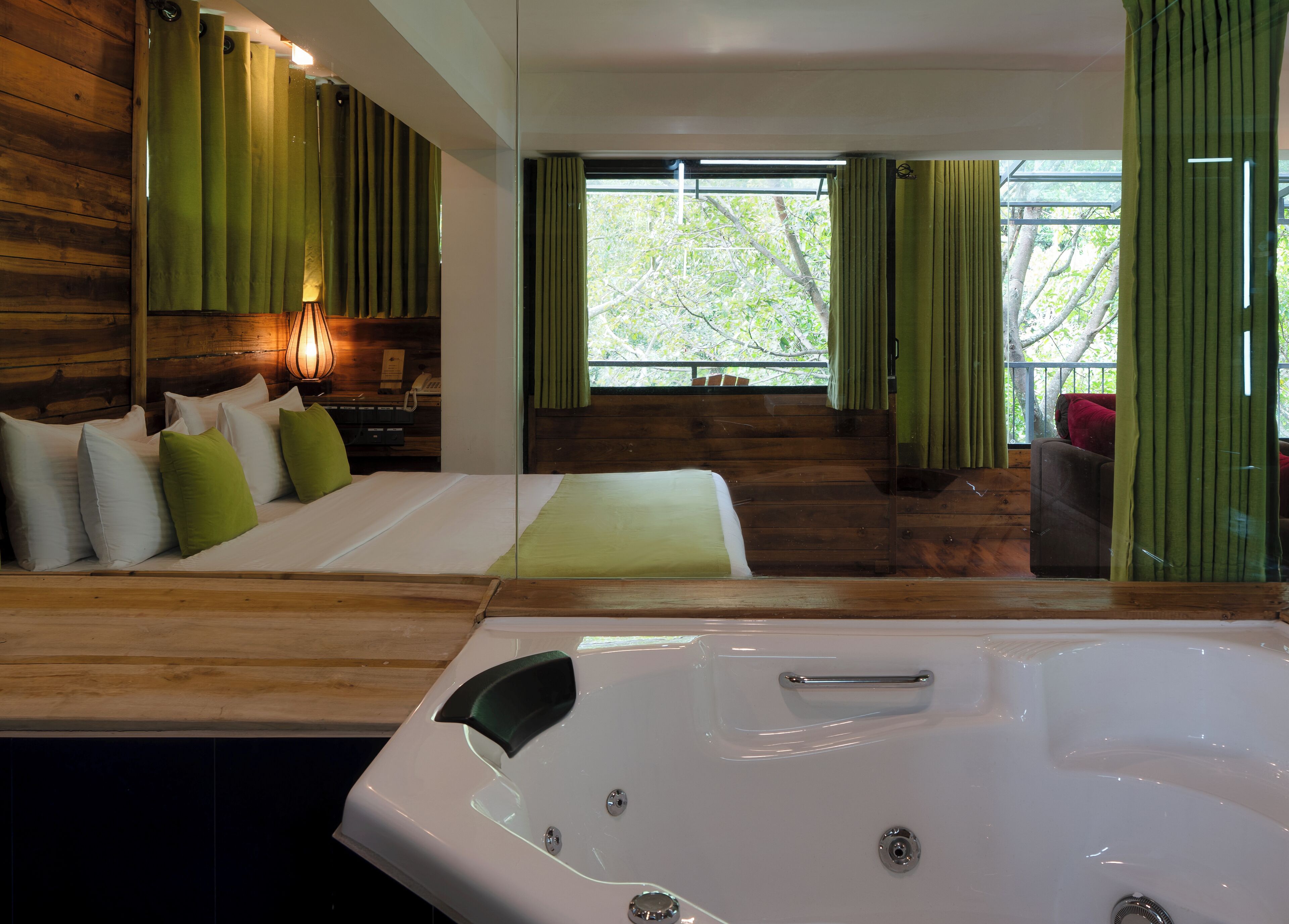 Ein modernes Zimmer mit Holzboden, einer großen Badewanne, einem Bett mit grün-weißen Kissen und Blick auf Bäume durch ein Fenster.