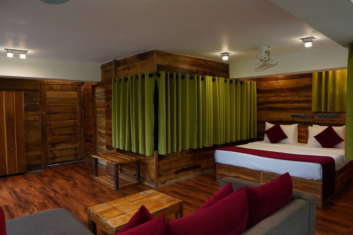 Quarto com cama de madeira, cortinas verdes, um banquinho de madeira e um sofá com almofadas vermelhas.
