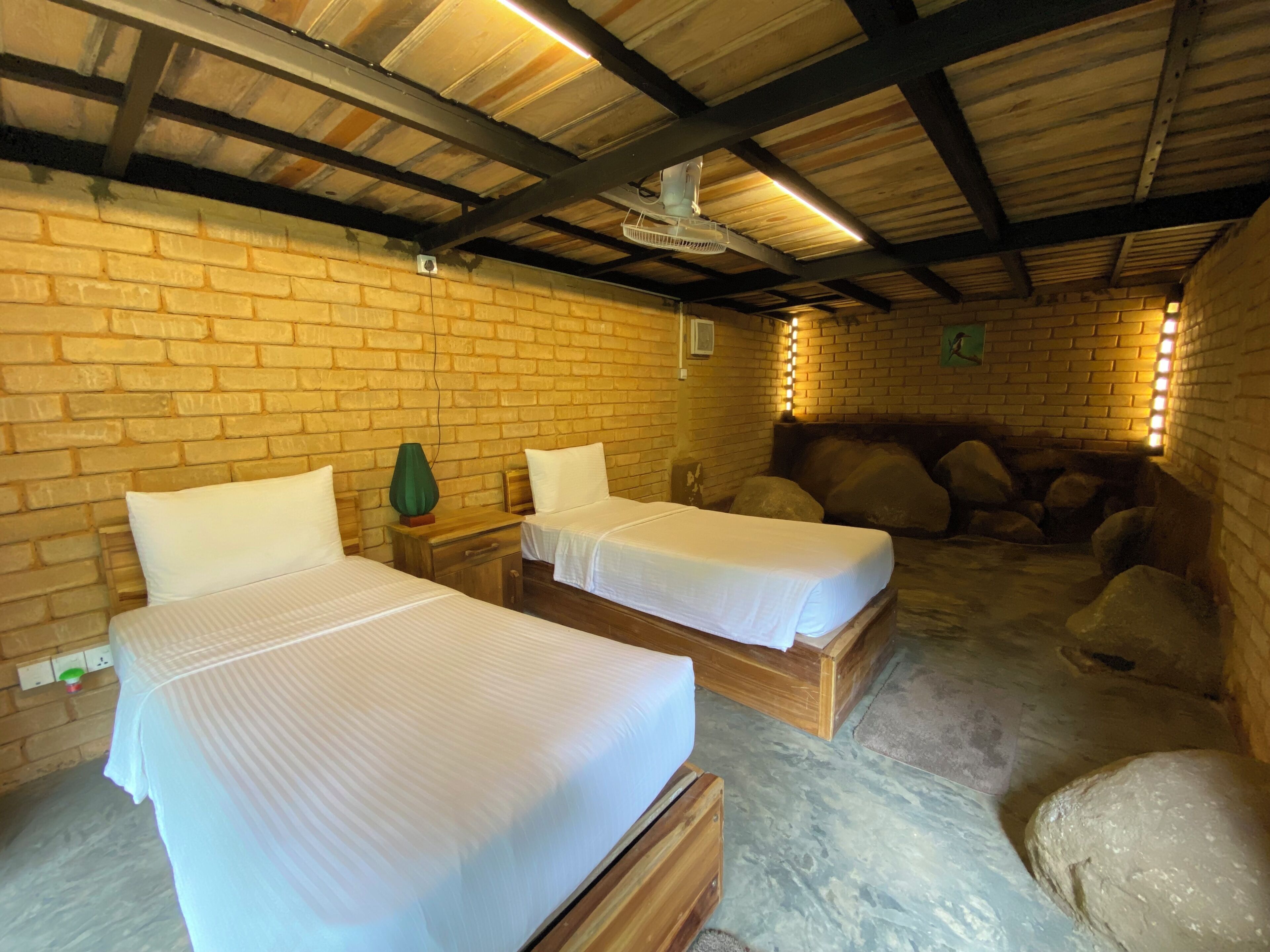 A room with two wooden beds, a brick wall, a ceiling fan, and a stone wall with a light fixture.