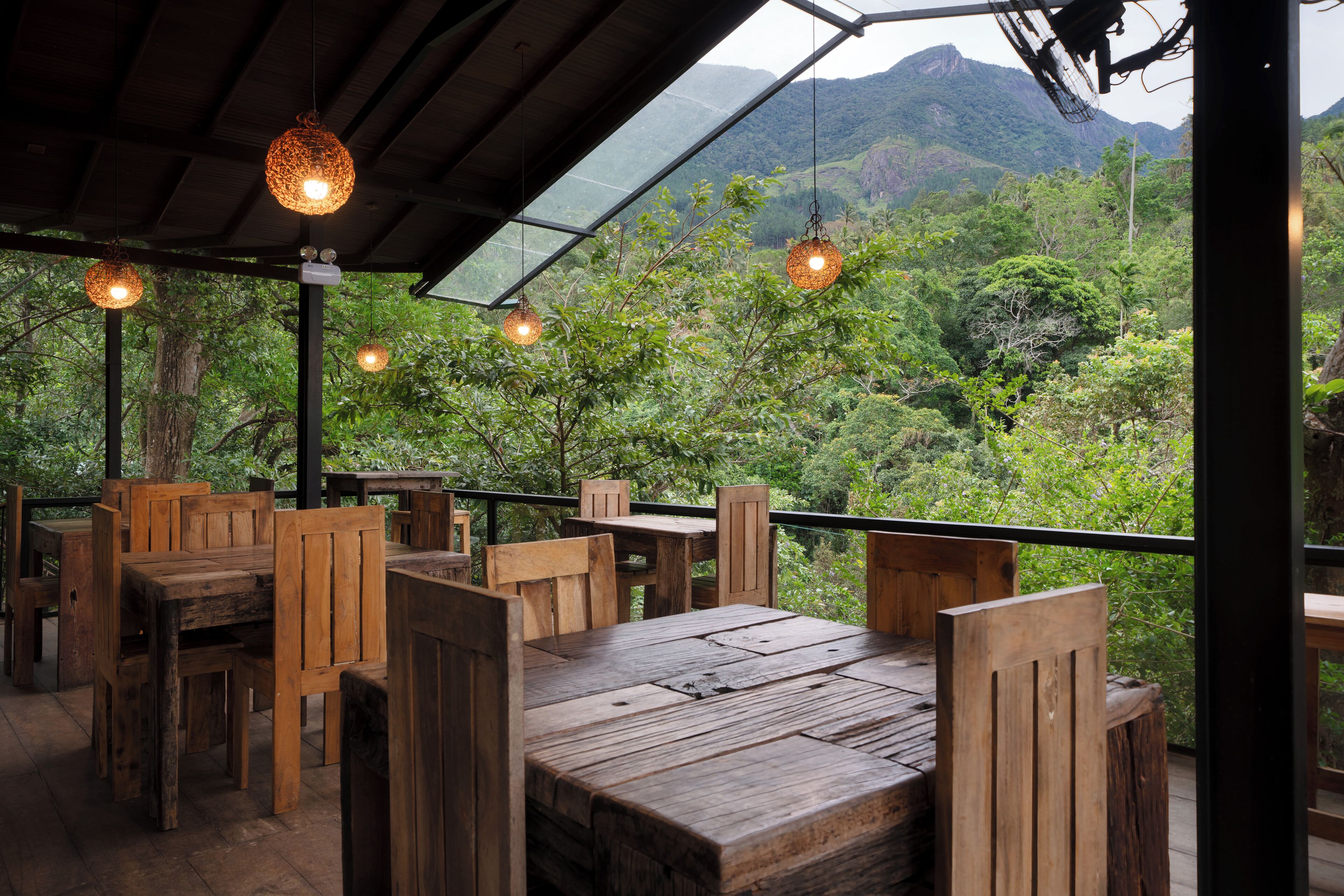 An outdoor dining area with wooden tables and chairs, hanging pendant lights, and a view of lush greenery and mountains in the background.