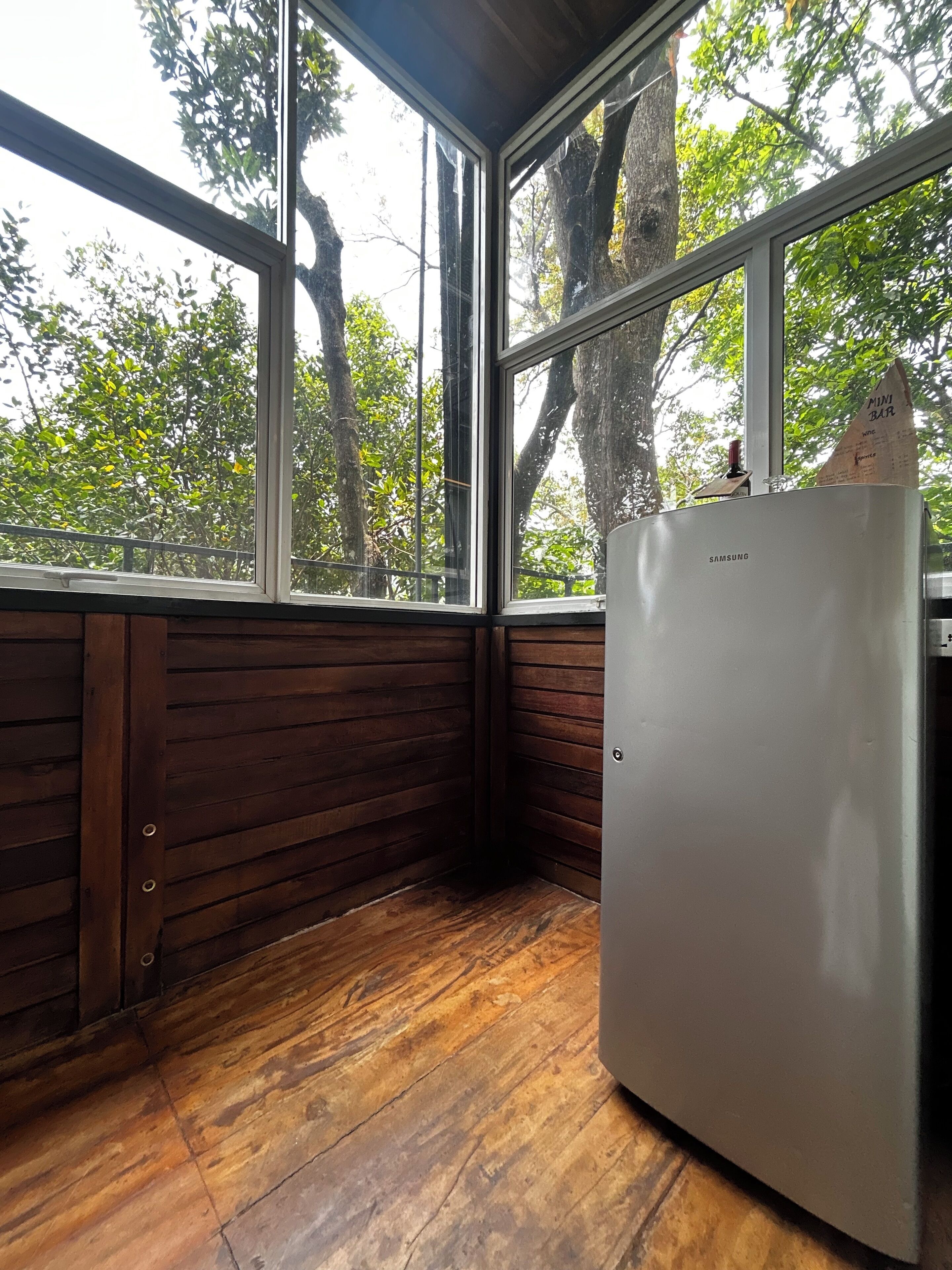 Une pièce d’angle aux murs en bois, avec un réfrigérateur Samsung blanc et de grandes fenêtres donnant sur la forêt.