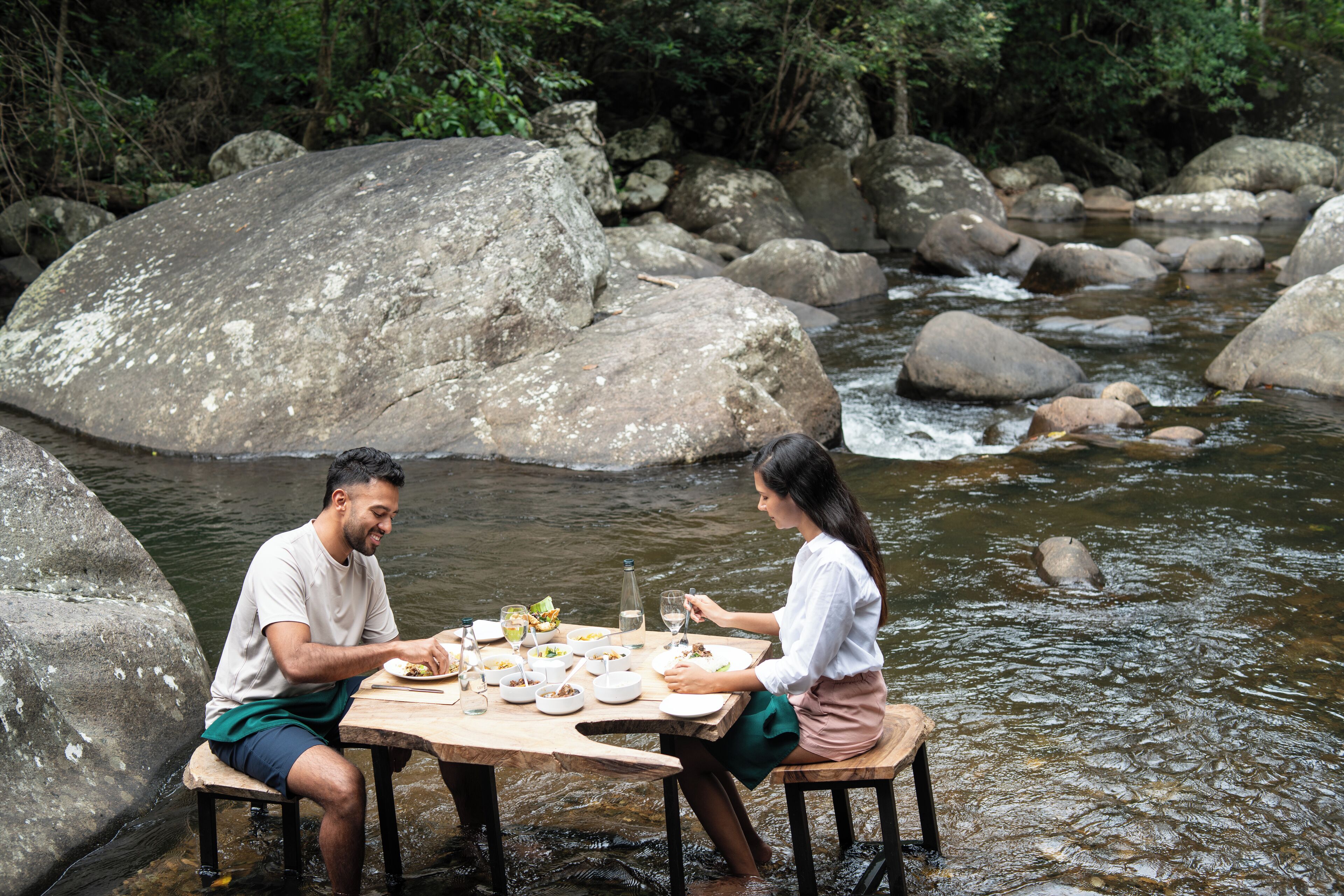 Mesa de jantar montada às margens de um rio, com pedras e vegetação ao fundo.