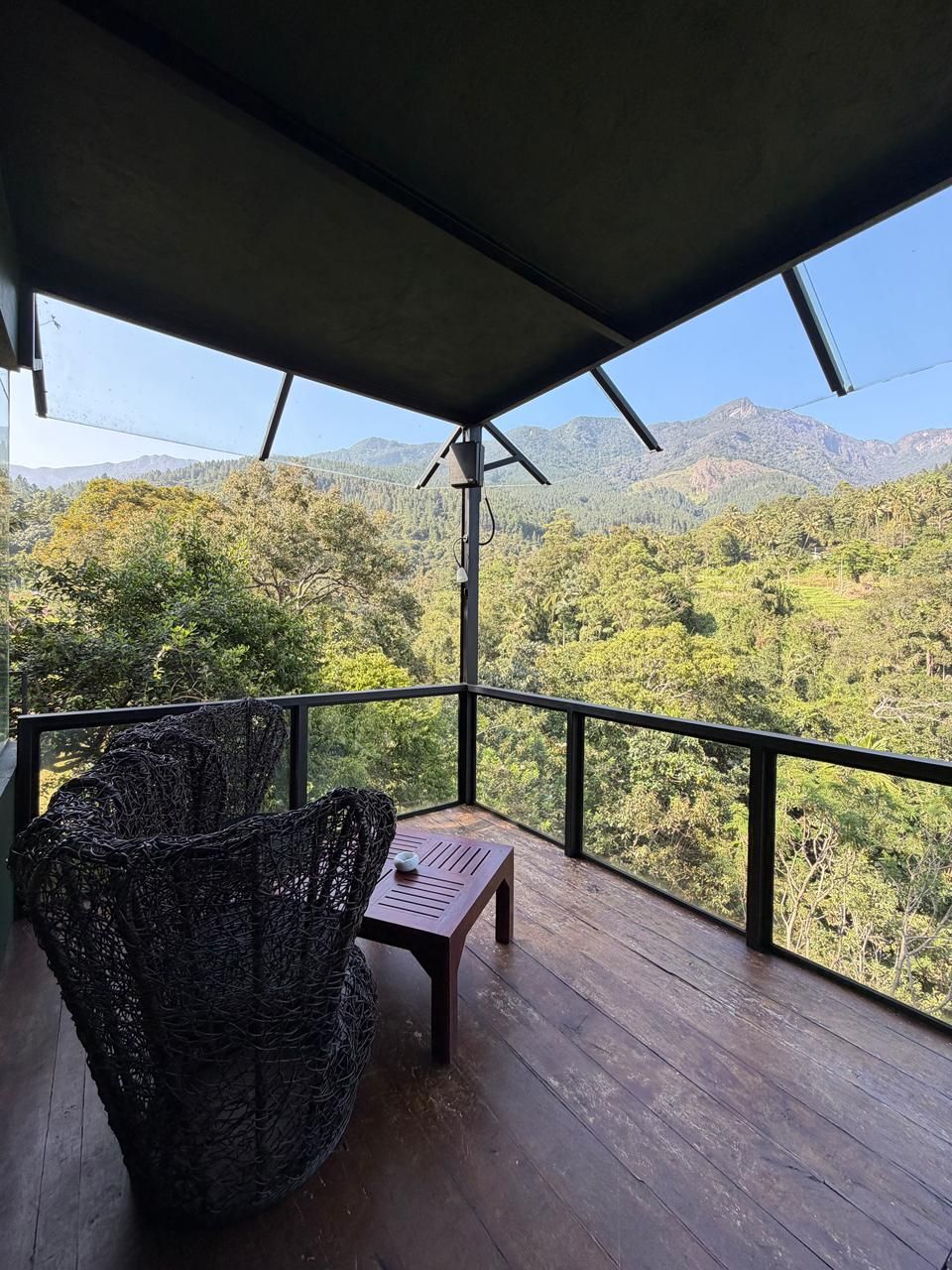 A balcony with a wicker chair and a small table, overlooking a mountainous landscape.