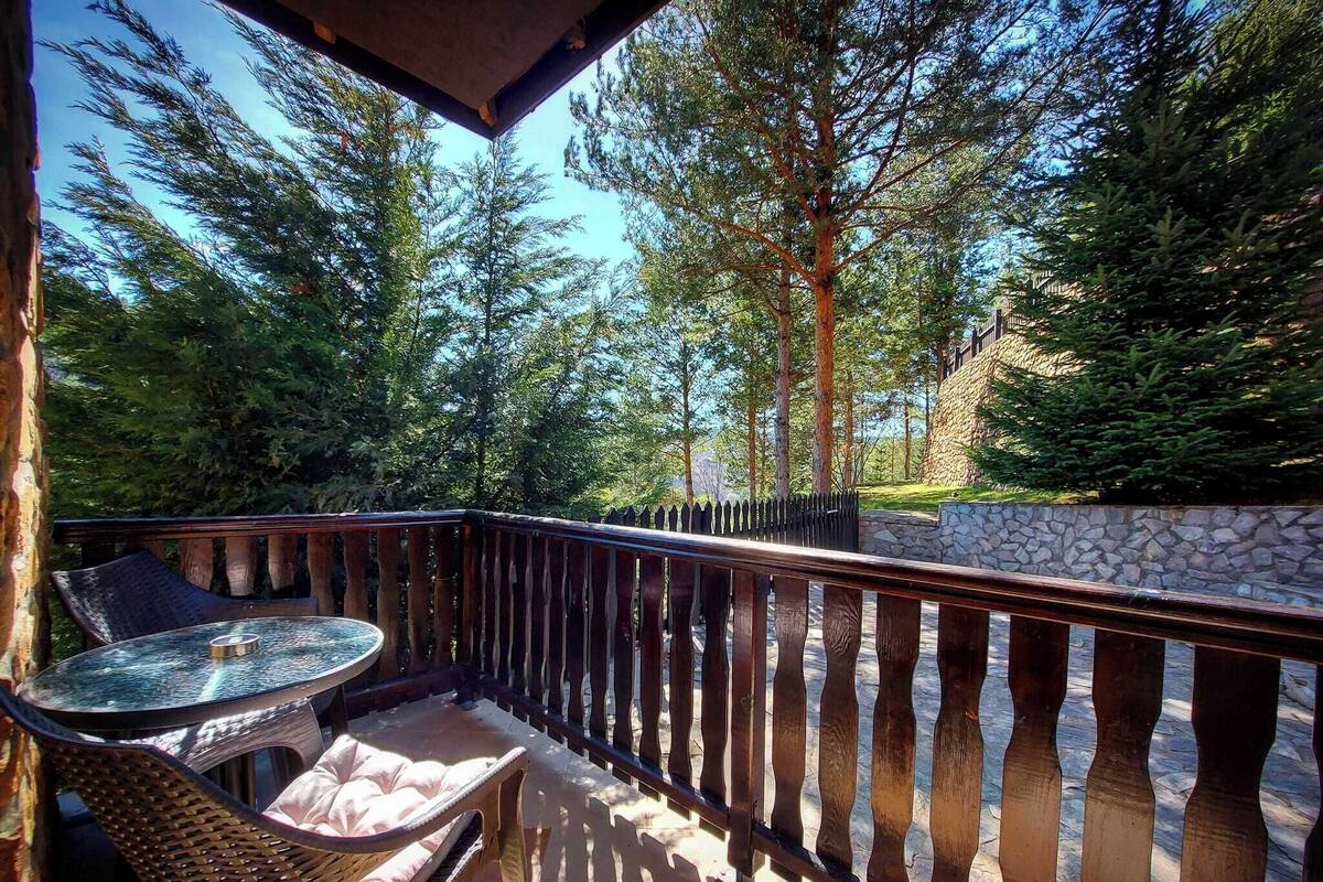 A balcony with a round table and chairs, surrounded by a wooden railing and stone wall, overlooking a forested area.