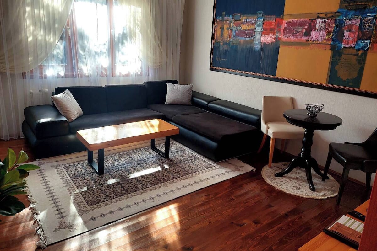 A living room with a black sofa, a wooden coffee table, a patterned rug, a round table with a bowl, and a large abstract painting on the wall.