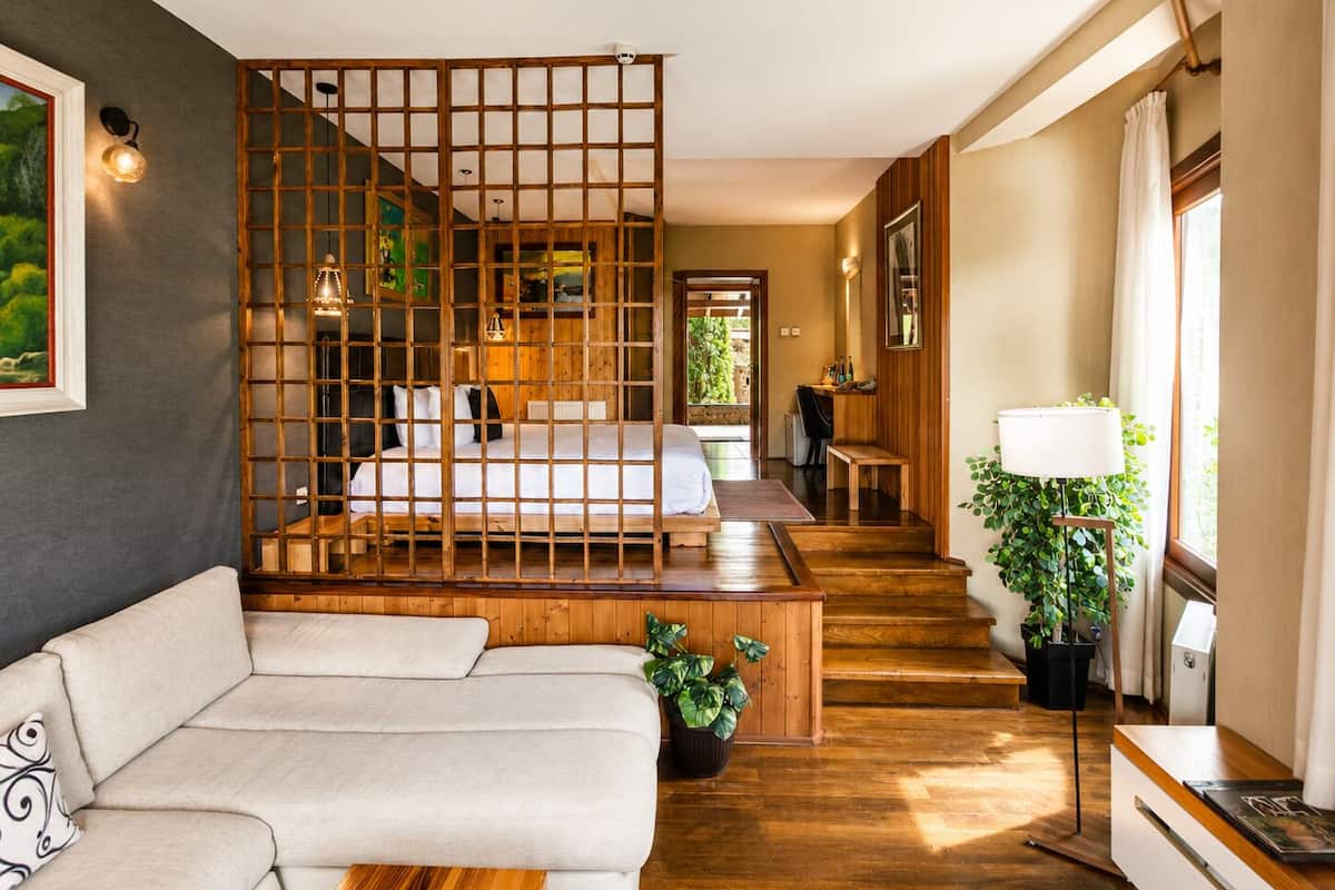A modern hotel room with a wooden partition, a sofa, a desk, a lamp, and a view of the garden.