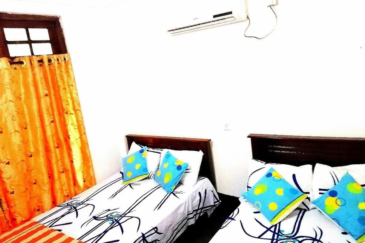 Une chambre avec deux lits simples, chacun doté d’une tête de lit en bois, d’une literie blanche à pois bleus et jaunes, et d’une fenêtre avec un rideau.