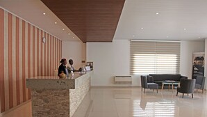 A reception area with a stone counter, two staff members, a sofa, and a window with blinds. - The Hub Hotel (Freetown)