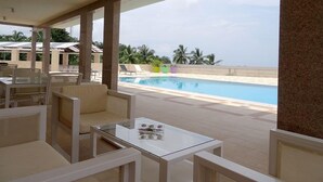 A poolside area with a glass table and chairs, a view of the pool, and palm trees in the background. - The Hub Hotel (Freetown)