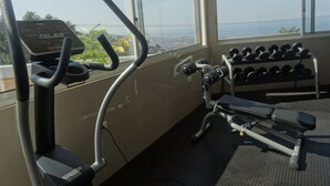 A gym with exercise equipment, including a treadmill and dumbbells, and large windows offering a view of the city and water. - The Hub Hotel (Freetown)