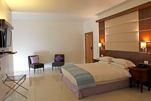A hotel room with a bed, two chairs, a TV, a desk, and a lamp. - The Hub Hotel (Freetown)