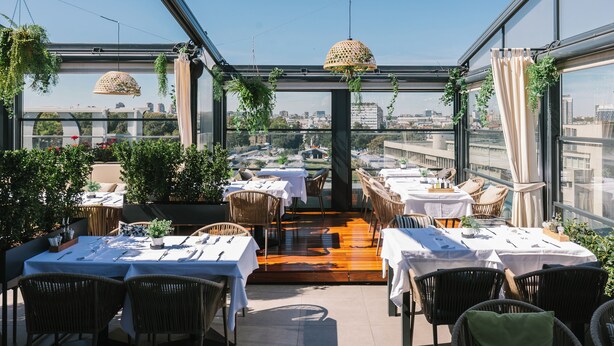 An outdoor dining area with tables and chairs, surrounded by greenery and large windows offering city views. - Hotel Heritage (Belgrade)