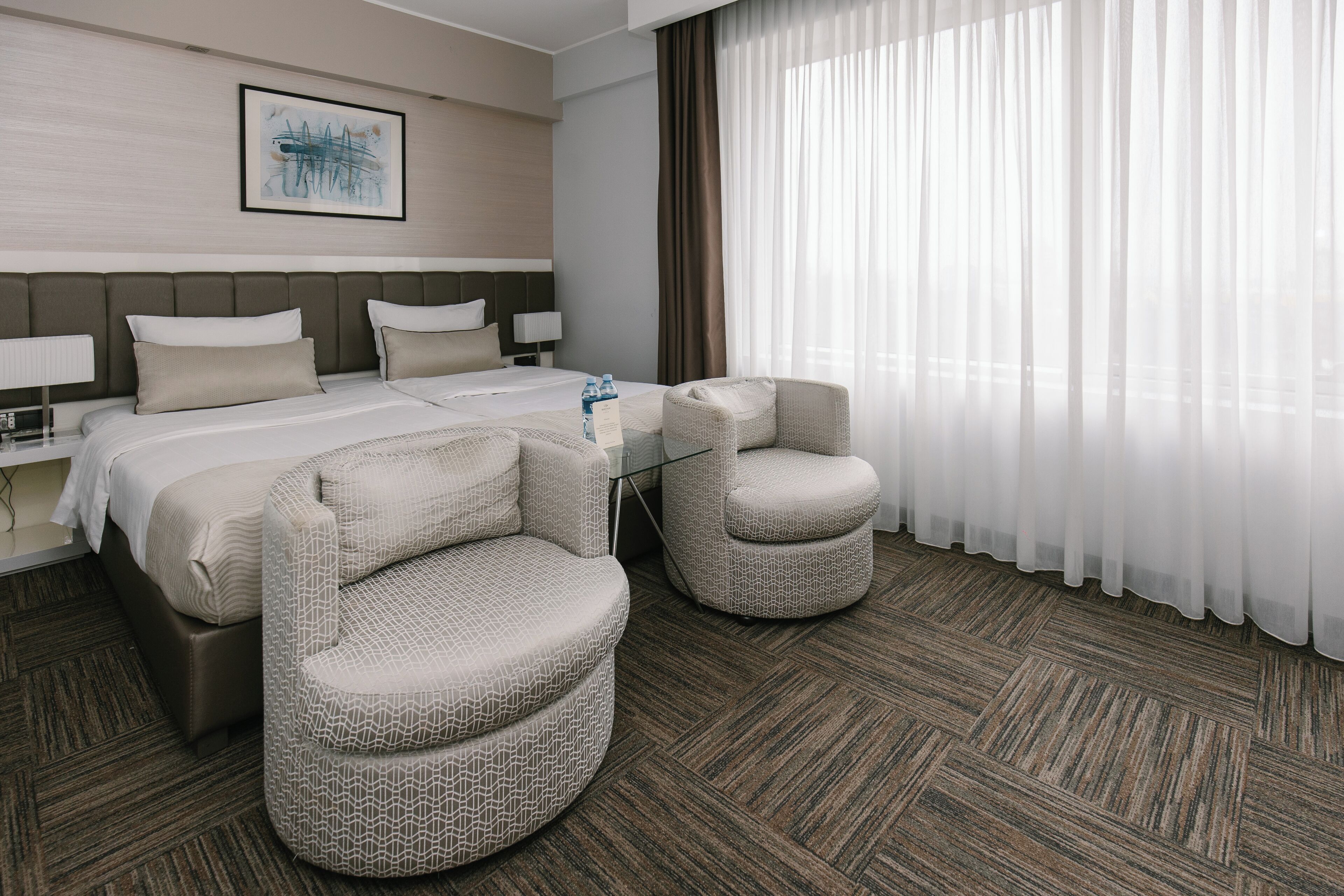 A modern hotel room with a large bed, two armchairs, a bedside table with a lamp, and a large window with sheer curtains.