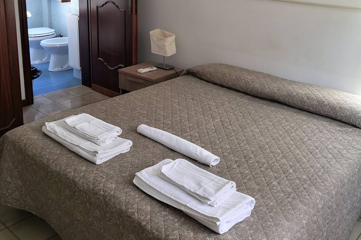 Una camera d'albergo con un letto, un quadro che raffigura un panorama urbano, un bagno con il water e il lavandino, e asciugamani sul letto.