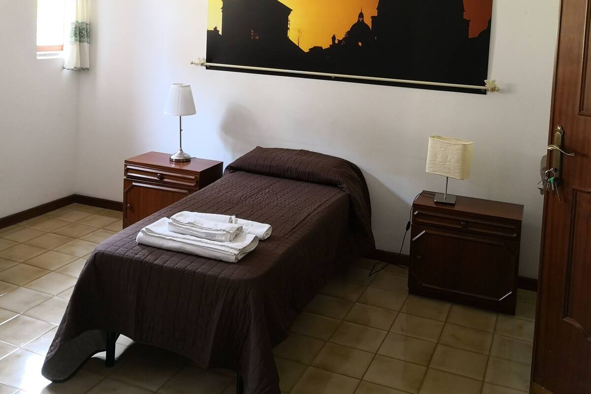 Una camera da letto con un letto, due comodini, una ventola a soffitto, una finestra con tende e un quadro appeso al muro raffigurante un paesaggio urbano.