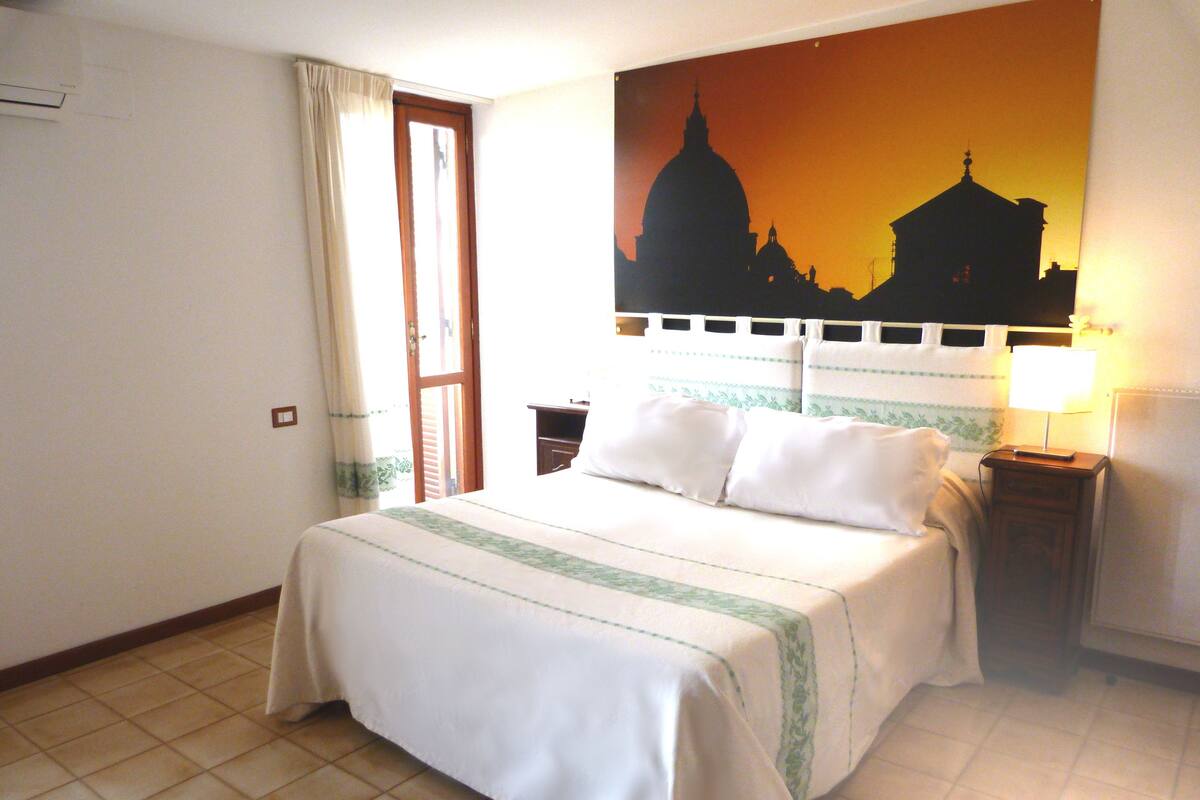 Una camera d'albergo con un letto, un comodino con una lampada, una finestra con tende e un quadro raffigurante un panorama cittadino.