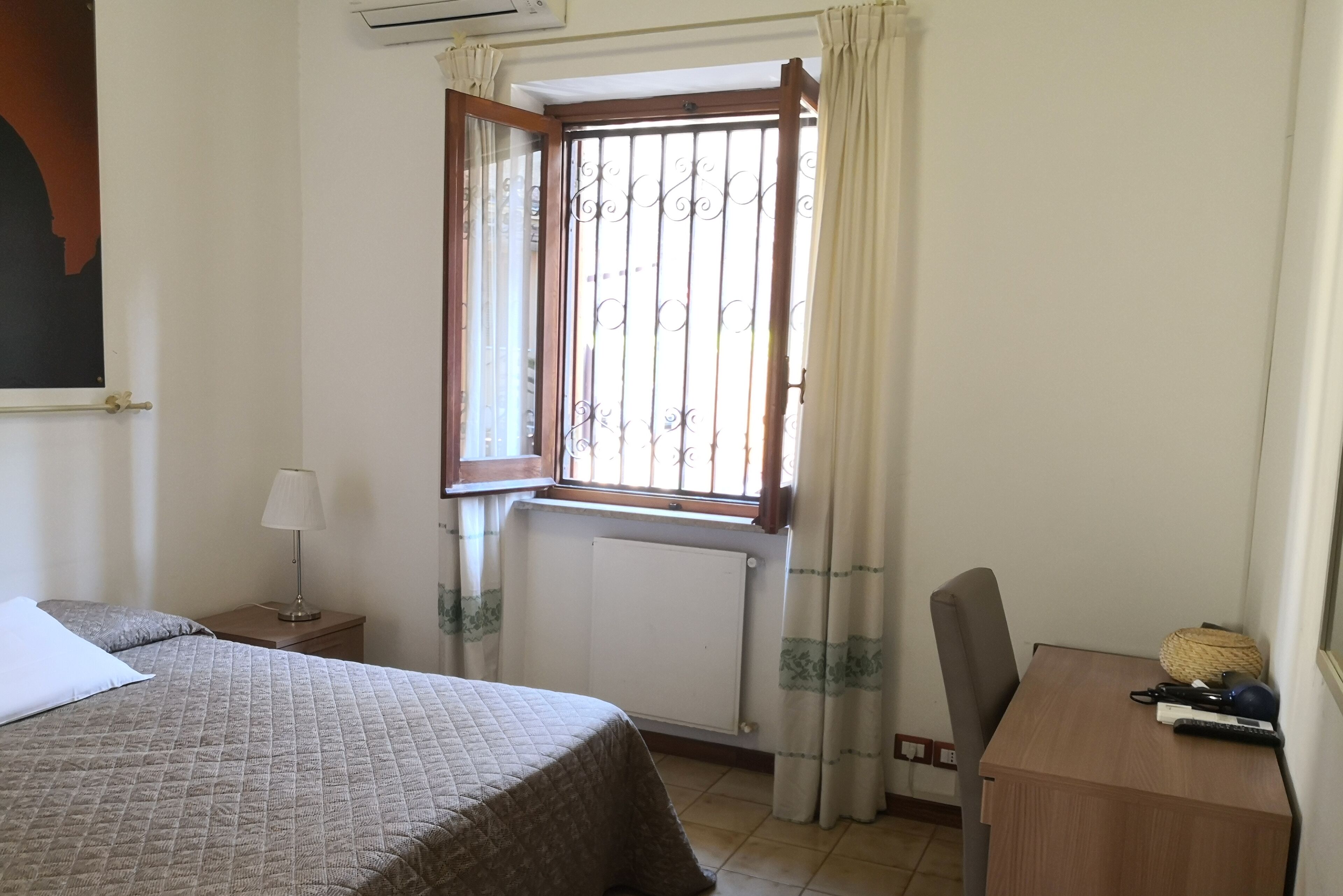 A bedroom with a bed, a nightstand, a desk, a chair, a window with curtains, and a radiator.