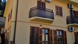 Un edificio de varios pisos con balcones, unidades de aire acondicionado y un patio empedrado con mesas al aire libre.