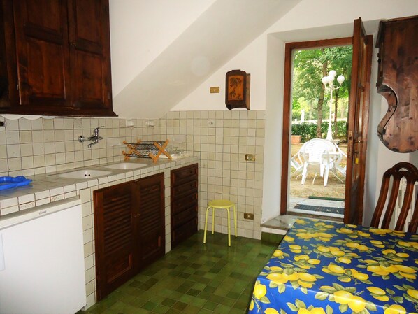 A compact kitchen with a sink, wooden cabinets, and a small stool. A dining area with a table and chairs is visible through an open door. - Agriturismo Villa Augusto (Cortona)
