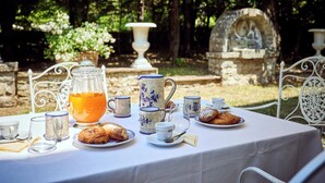 An outdoor dining table set with tea, coffee, and pastries, surrounded by white metal chairs and a stone fountain in the background. - Agriturismo Villa Augusto (Cortona)