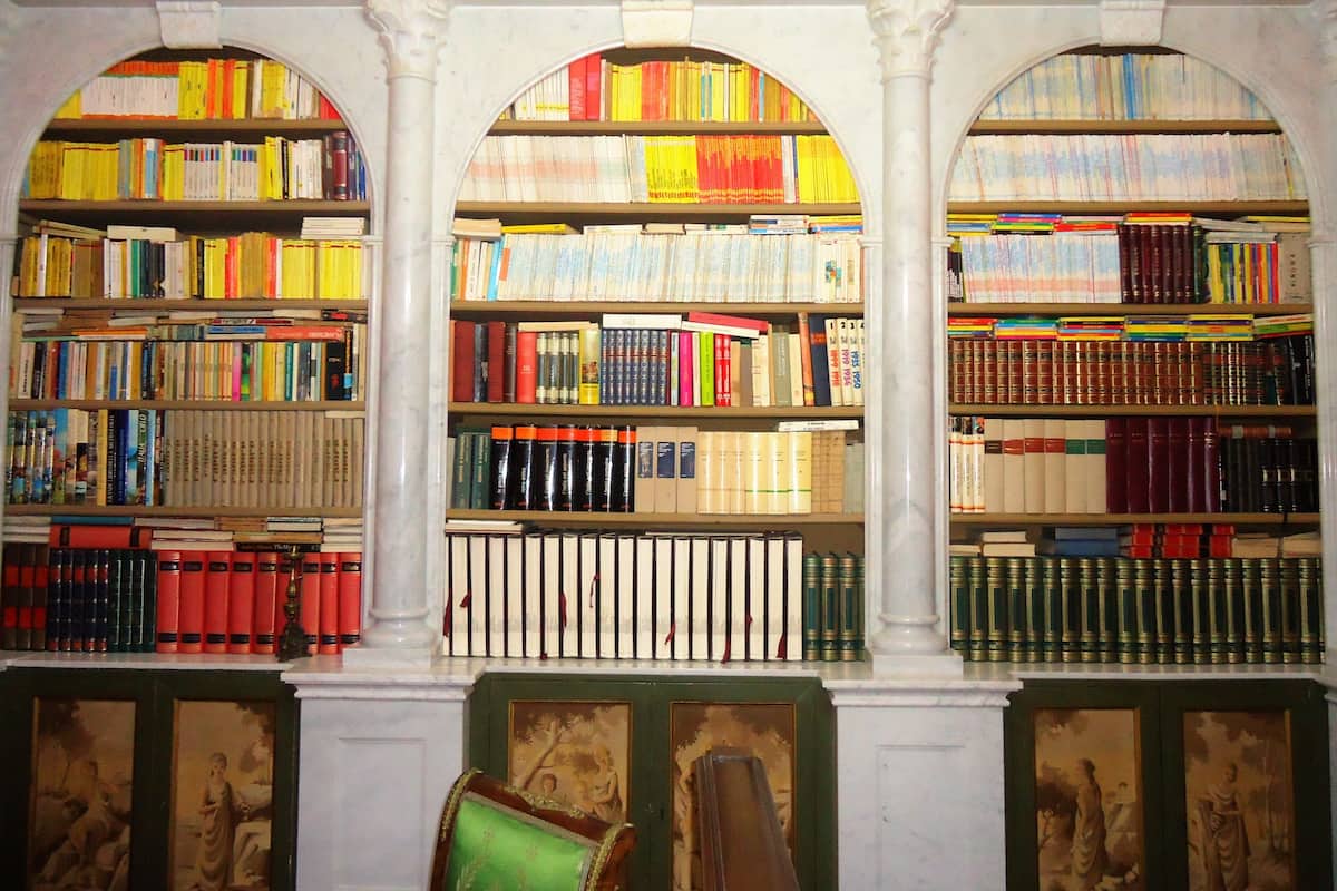 A library with bookshelves filled with books, framed artwork on the walls, and a green chair in the foreground.