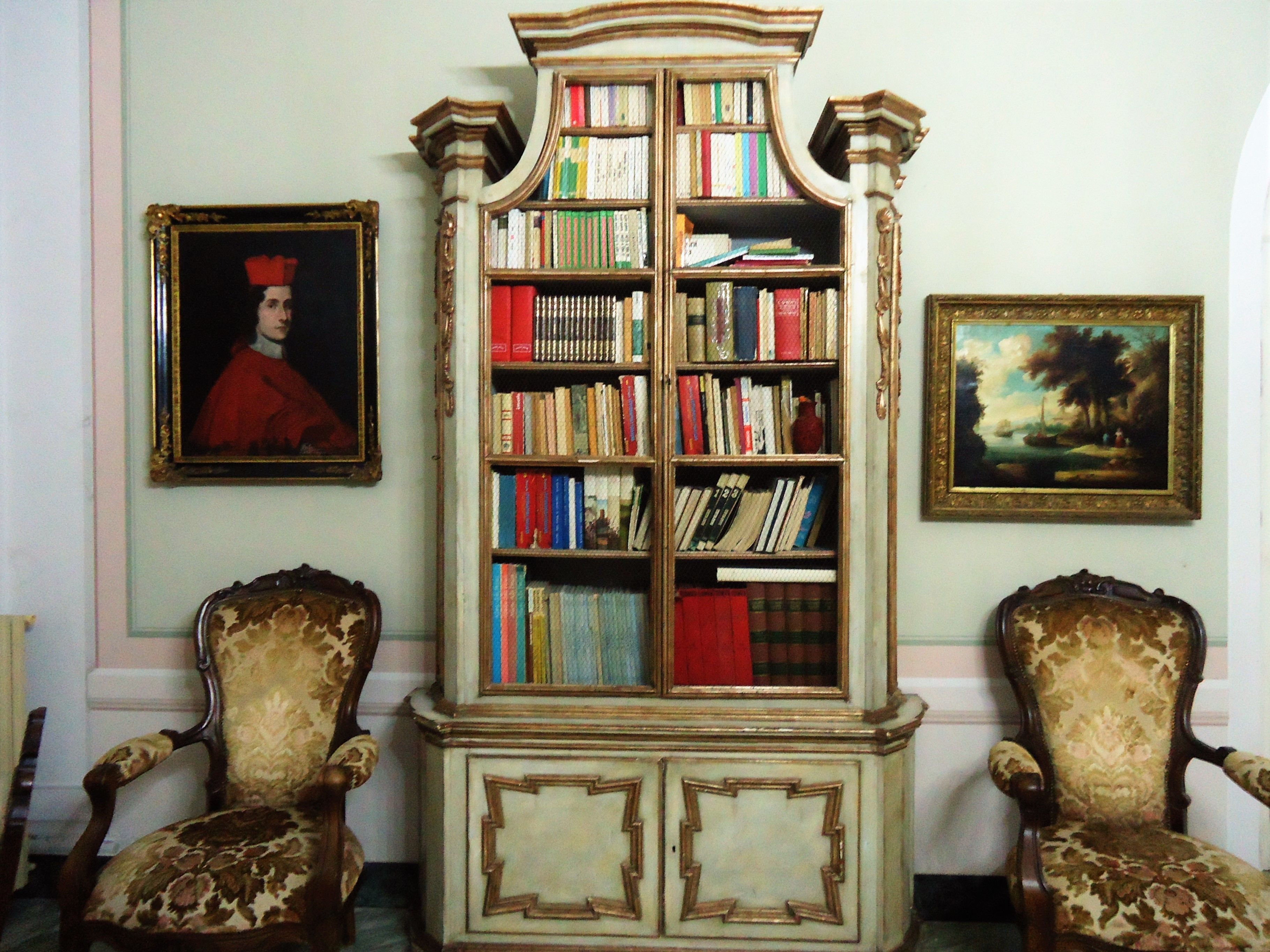 Ein Zimmer mit einem großen Bücherregal, zwei verzierten Stühlen, einem gerahmten Porträt und einem Landschaftsbild an der Wand.