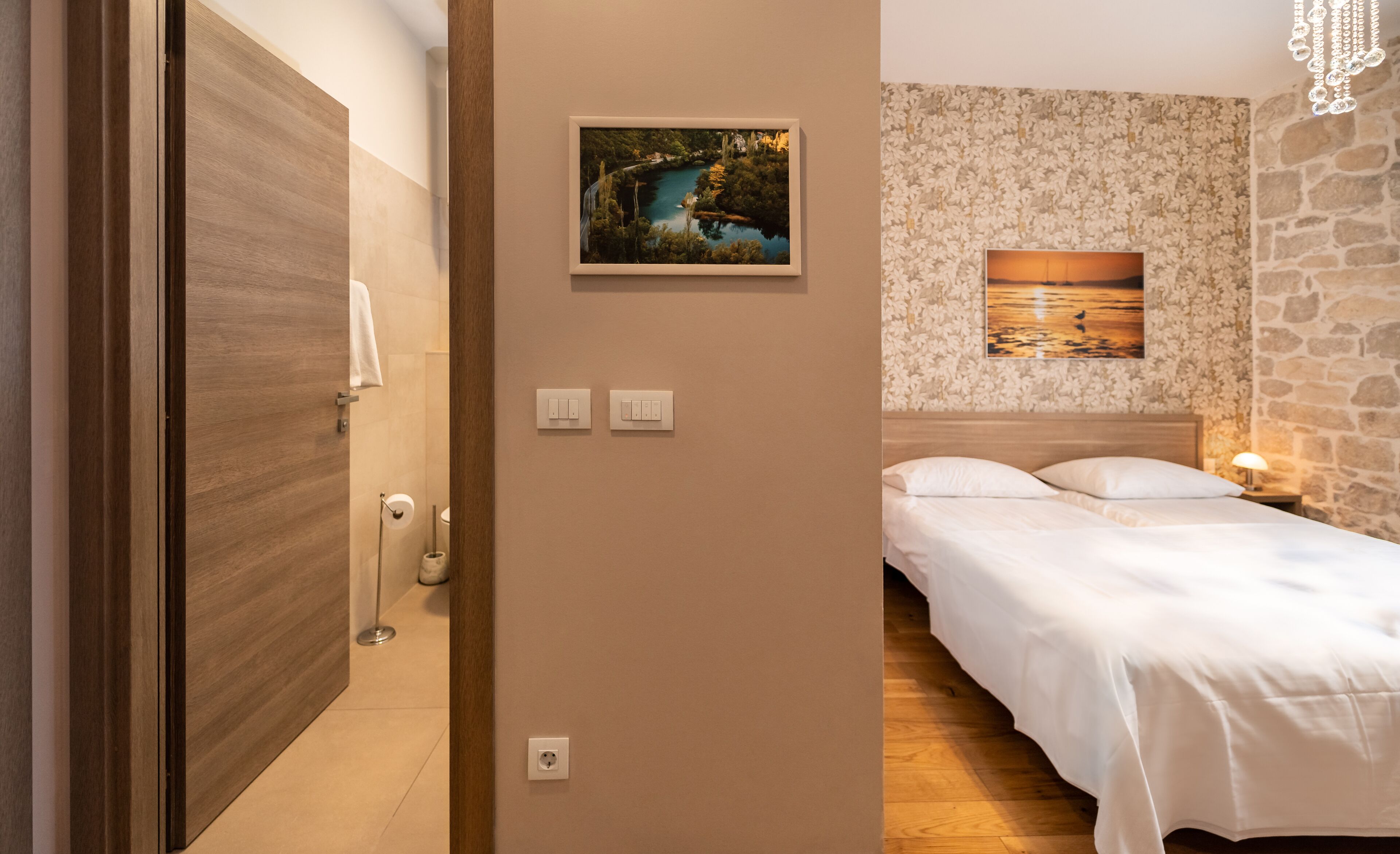 A hotel room with a wooden door, a bathroom with a toilet and sink, a bed with white bedding, and two wall-mounted light switches.