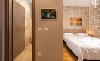 Une chambre d’hôtel avec une porte en bois, une salle de bain avec des toilettes et un lavabo, un lit avec du linge de lit blanc, et deux interrupteurs fixés au mur.