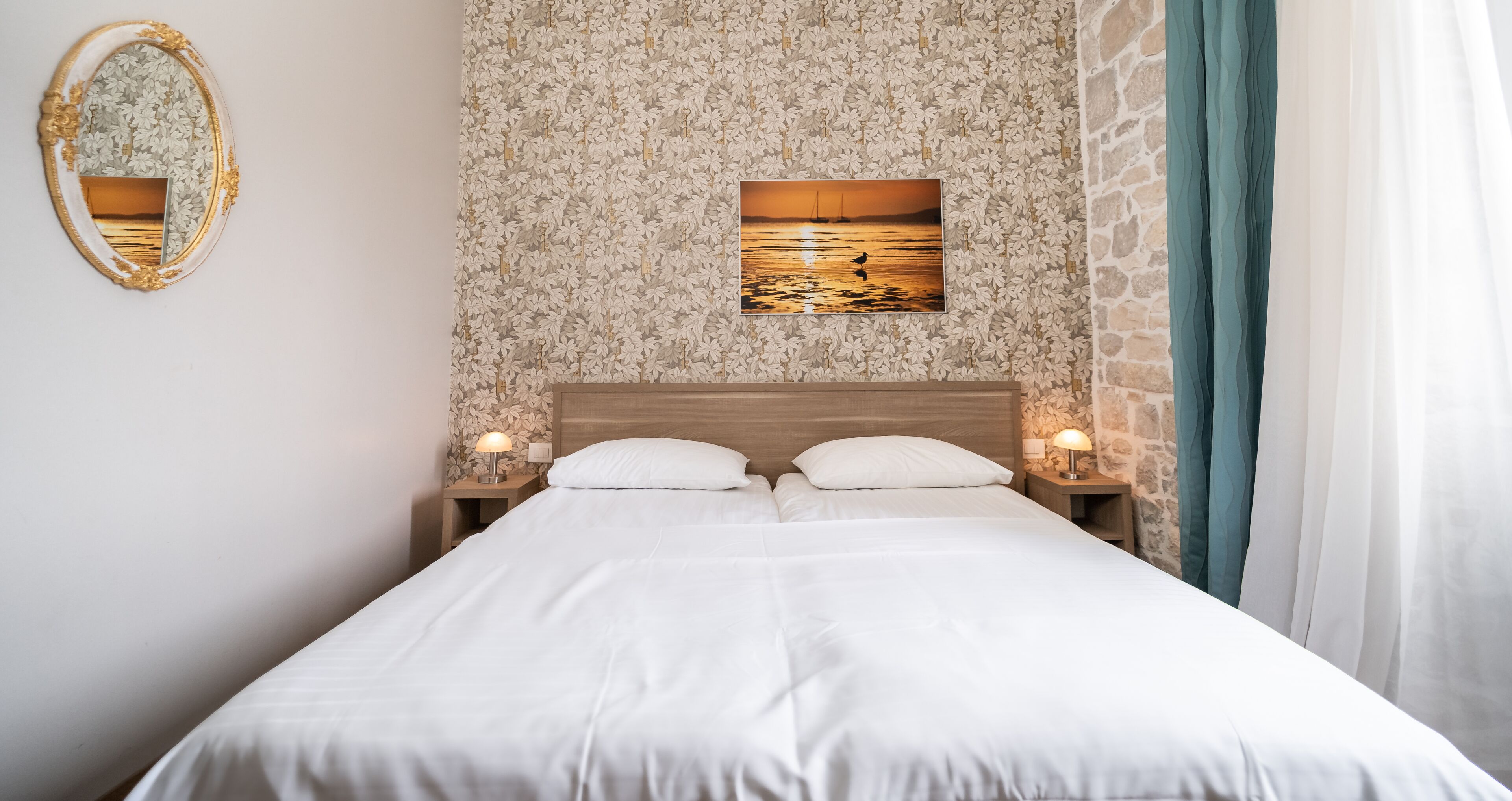 A neatly made bed with white linens, a wooden headboard, and a floral-patterned wallpaper. There is a framed picture of a sunset on the wall, and a mirror with a gold frame on the left.