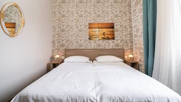 A neatly made bed with white linens, a wooden headboard, and a floral-patterned wallpaper. There is a framed picture of a sunset on the wall, and a mirror with a gold frame on the left.