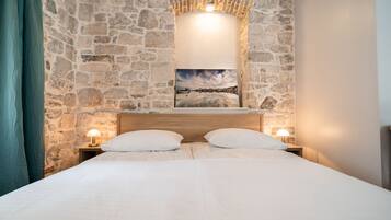 A bedroom with a stone wall, a wooden headboard, a bed with white linens, two bedside lamps, and a framed picture above the bed.