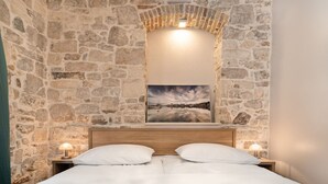 A bedroom with stone walls, a wooden headboard, two white pillows, a nightstand with a lamp, and a framed picture of a coastal scene. - Tifani Luxury Rooms 2 (Split)