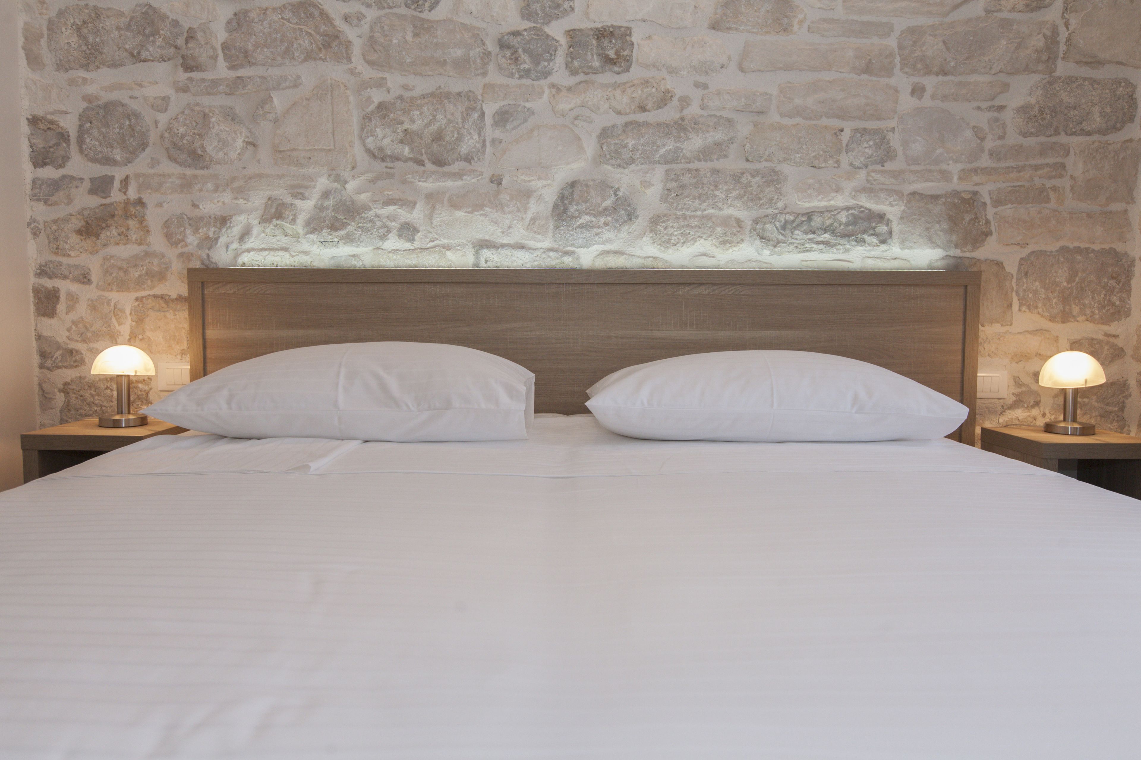 Cama con ropa de cama blanca y dos almohadas, flanqueada por mesitas de noche con lámparas, sobre un fondo de pared de piedra.