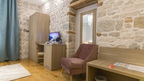 A room with a stone wall, a wooden desk, a pink chair, a wooden bench, and a television on a cabinet. - Tifani Luxury Rooms 2 (Split)