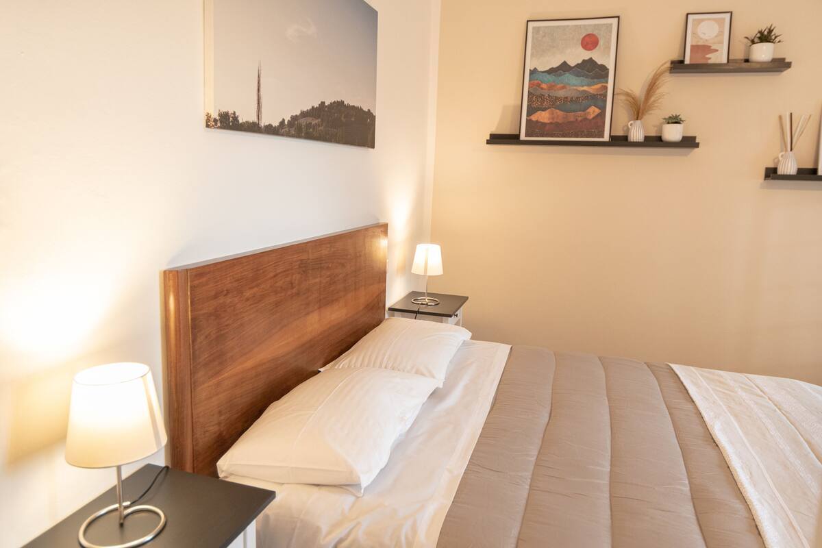 Uma cama bem arrumada com lençóis brancos, cabeceira de madeira, luminária de cabeceira e mesa de cabeceira com abajur. Estantes fixadas na parede exibem objetos decorativos e quadros.