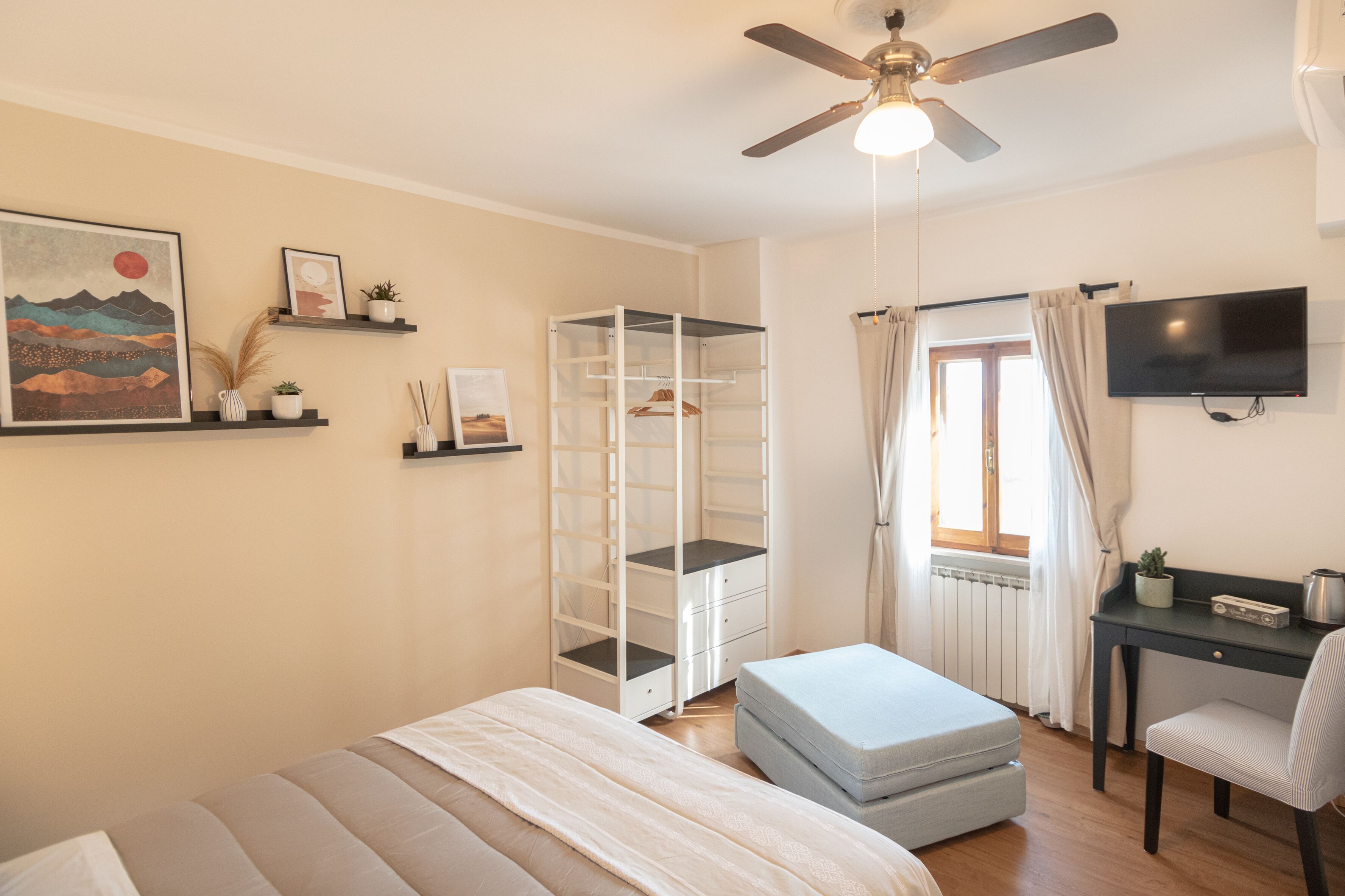 A bedroom with a bed, a desk, a chair, a ceiling fan, a TV, and a window with curtains.