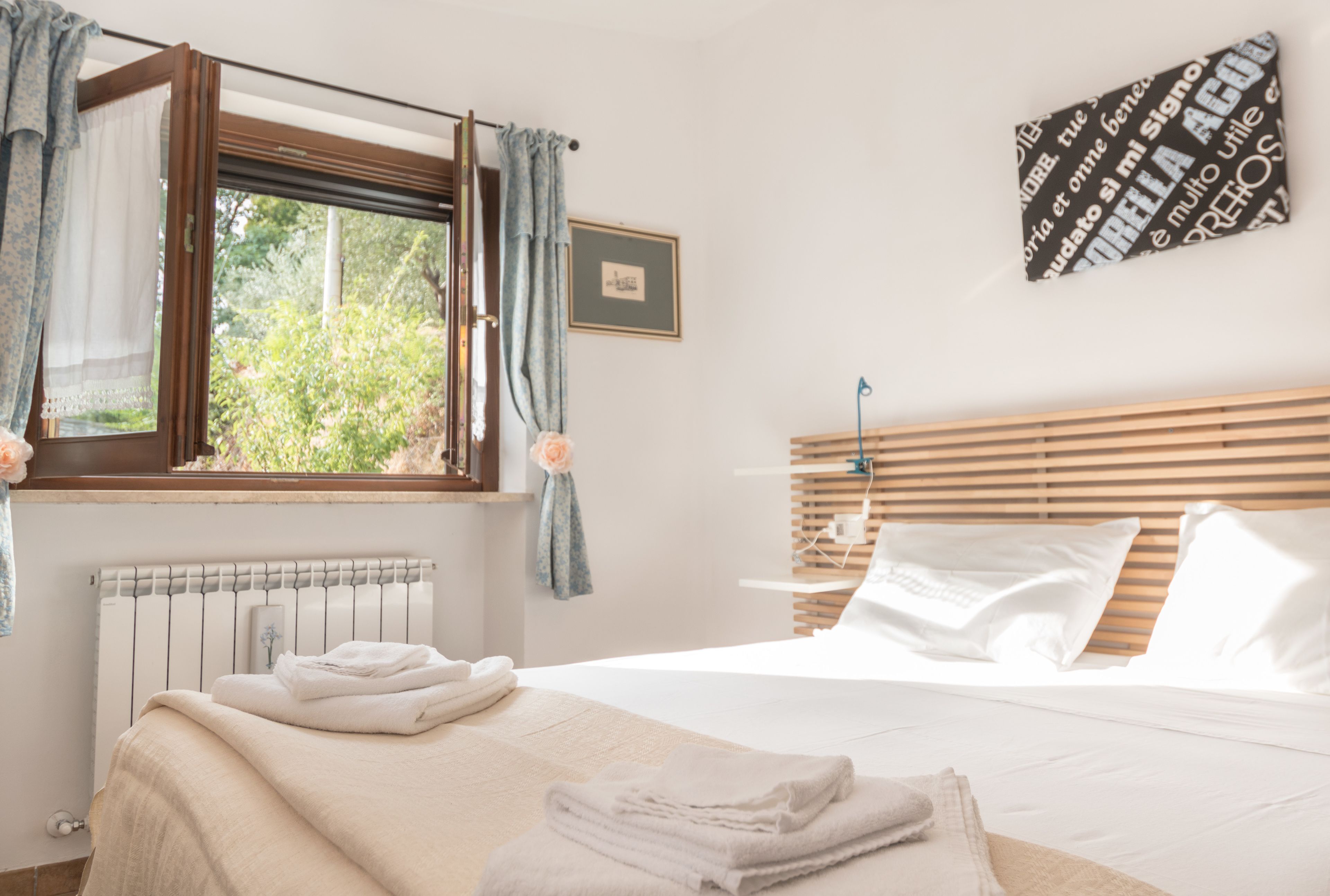 Una cama bien hecha con toallas dobladas, una ventana con cortinas, un radiador y una pared con un cuadro enmarcado.
