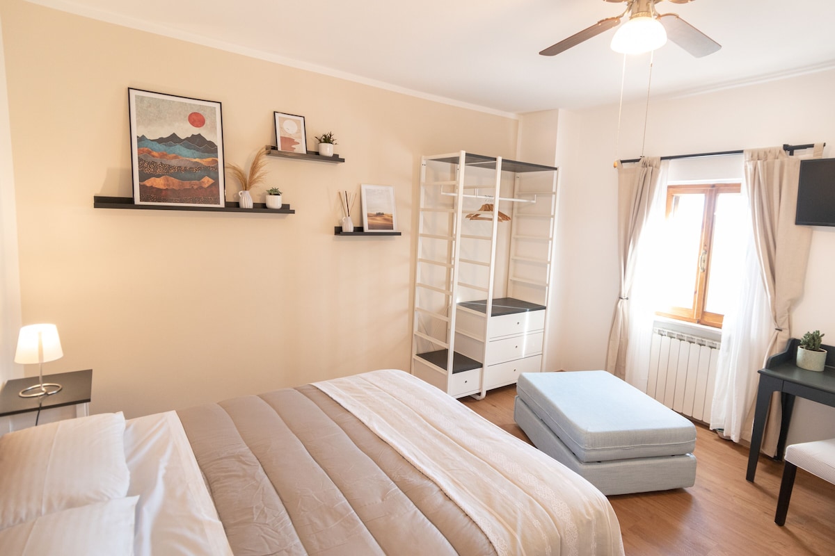 Une chambre à coucher comprenant un lit, une table de chevet, une commode, un ventilateur de plafond, une fenêtre avec des rideaux, un radiateur, une étagère avec des objets décoratifs et un téléviseur fixé au mur.