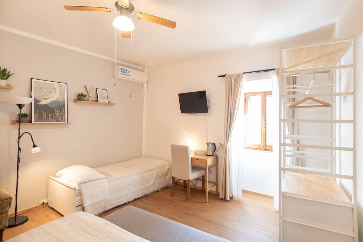 Quarto compacto com uma cama de solteiro, escrivaninha com cadeira, ventilador de teto, TV fixada na parede e janela com cortinas.