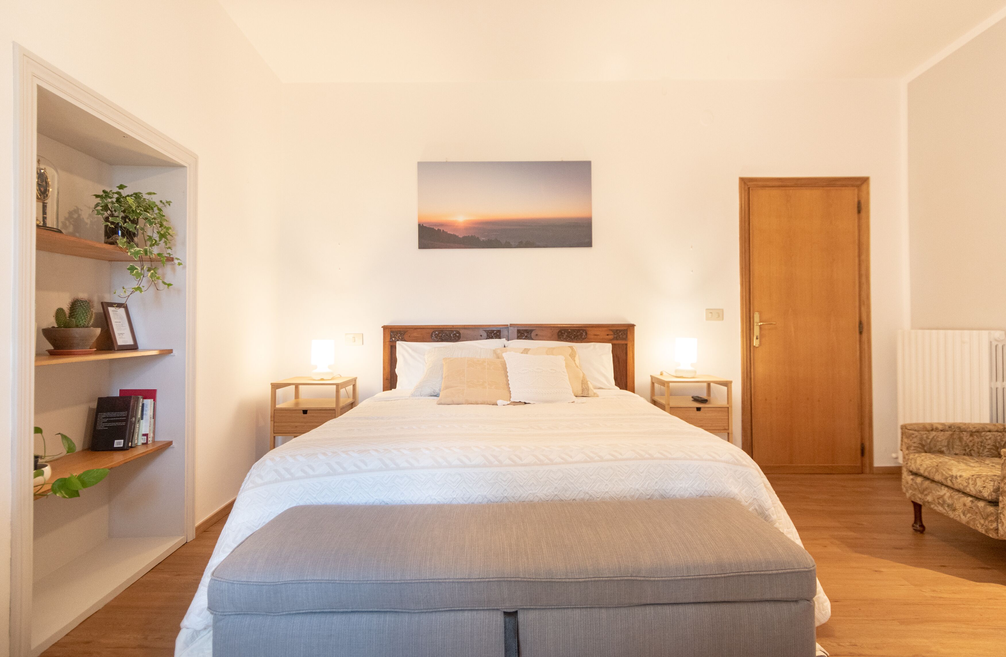 A bedroom with a large bed, two bedside tables, a wooden door, and a shelf with plants and books.