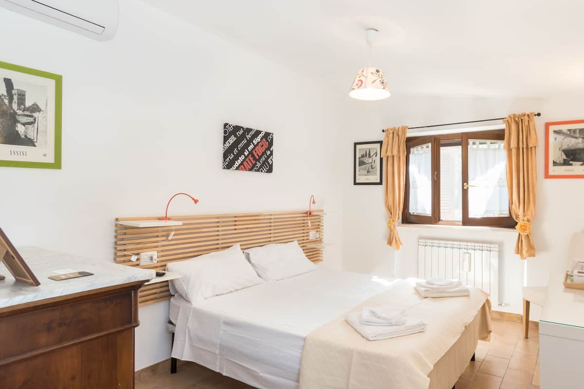 Quarto limpo e bem iluminado com cama, mesa de cabeceira e janela com cortinas.