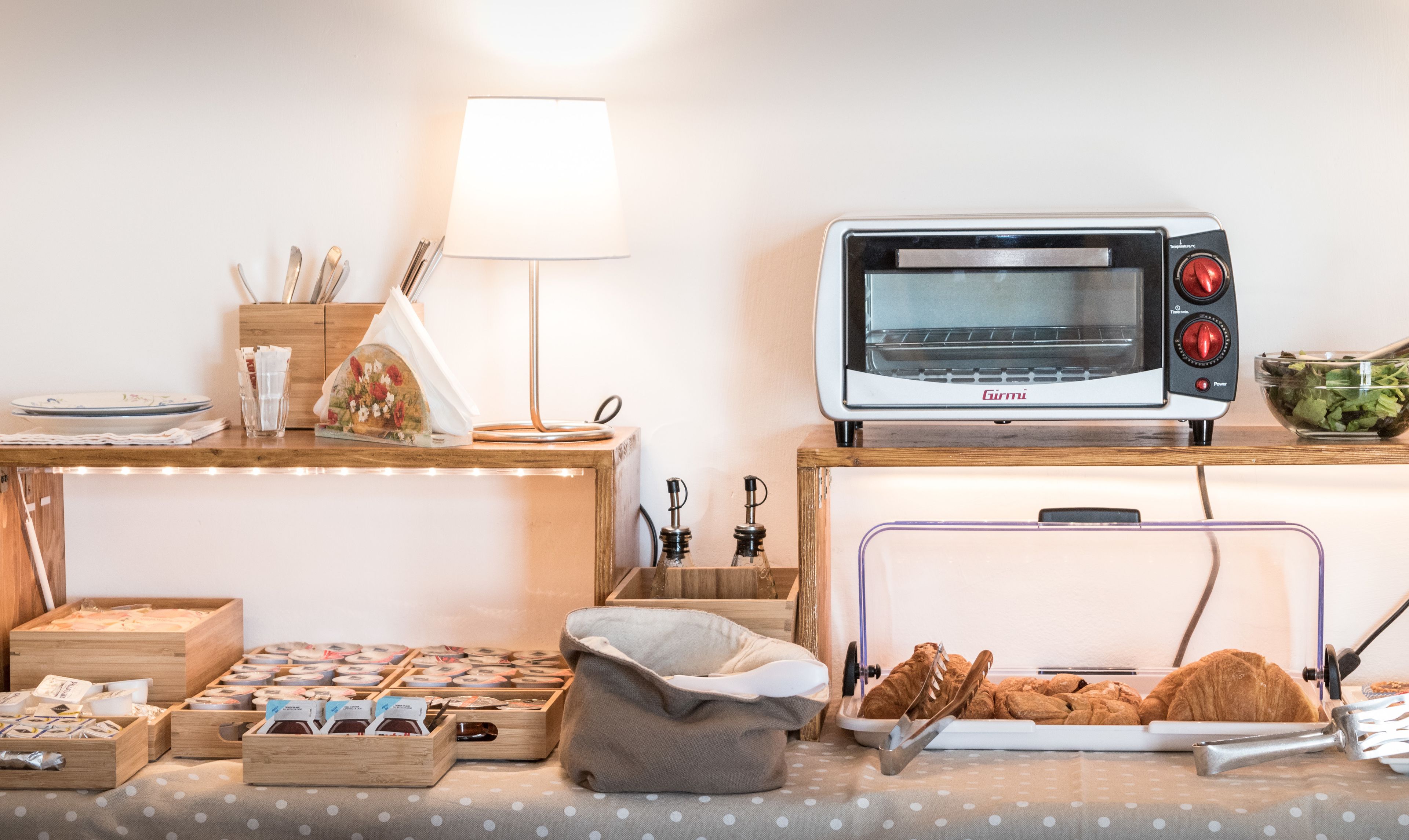 A kitchen with a Girmi toaster oven, a wooden table with a lamp, and various kitchen items.