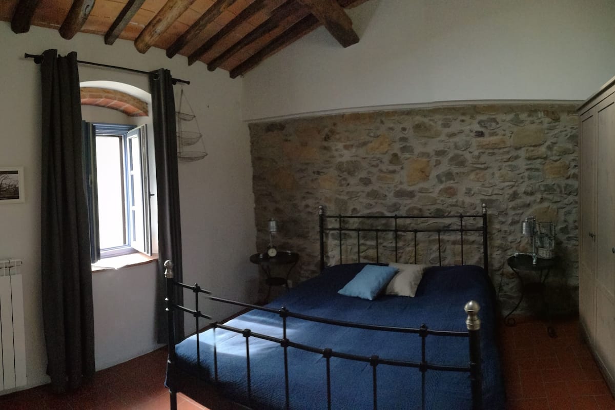 A bedroom with a stone wall, a bed with blue bedding, a radiator, and a window with curtains.