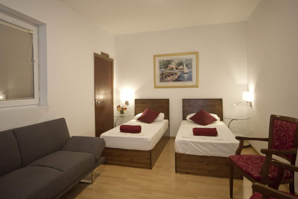 Et hotelværelse med to enkeltsenge, en sofa, en stol, et lille bord med en vase blomster og et billede på væggen.