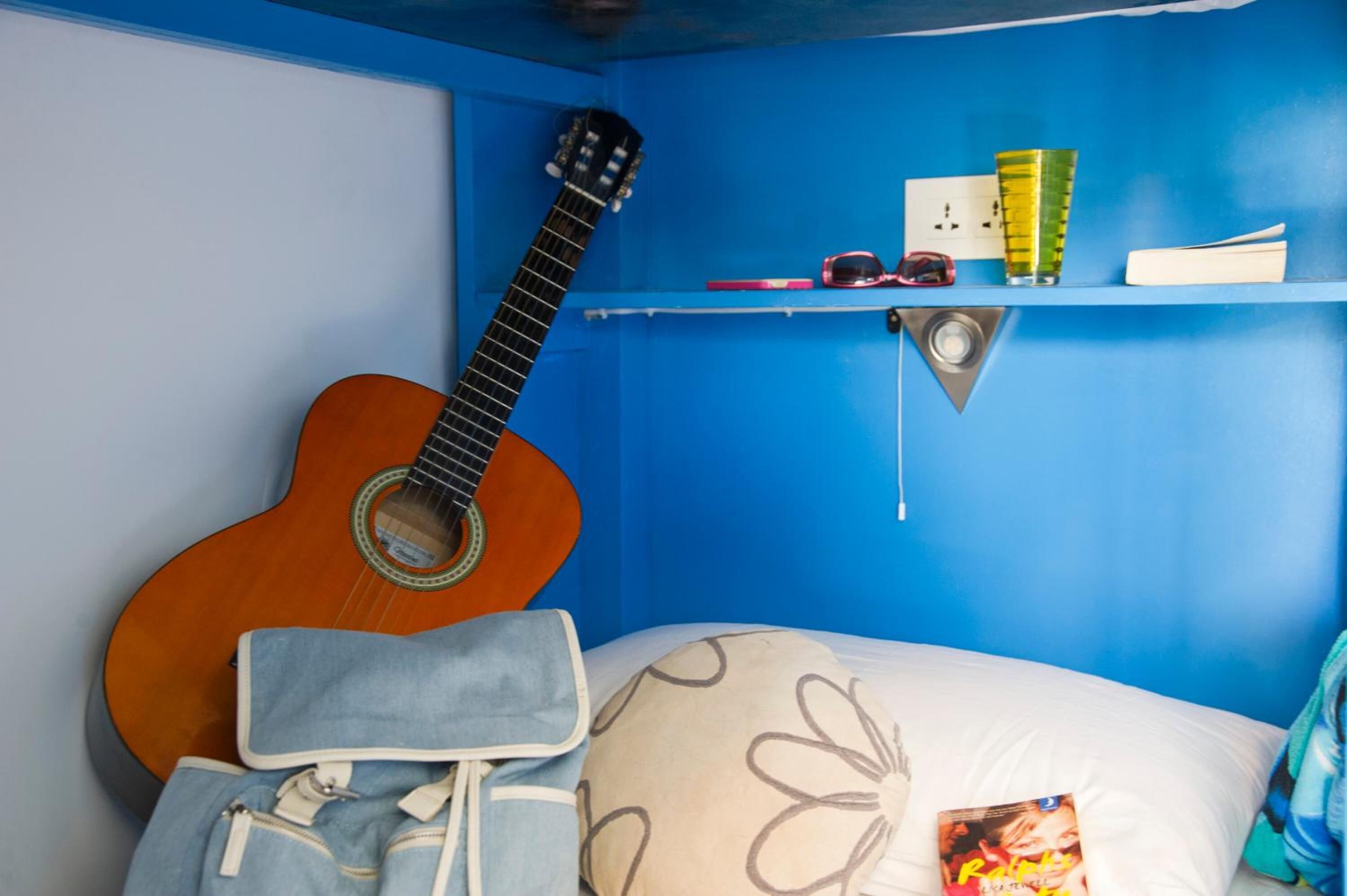 Une chambre avec une guitare, une étagère remplie de livres et de verres, et un lit avec un oreiller et une couverture.