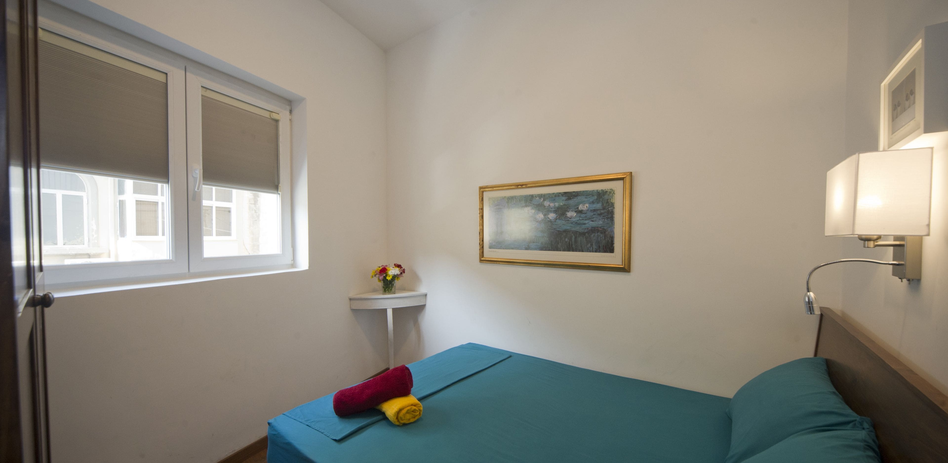 Ein kleiner, ordentlicher Raum mit einem Einzelbett, einem Fenster, einer Wandlampe und einem gerahmten Bild.