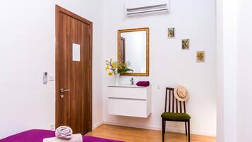 A room with a wooden door, a white vanity with a mirror, a small chair, and a purple bed.