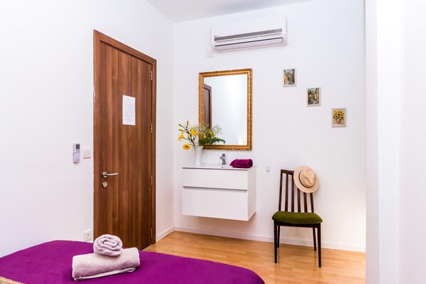A room with a wooden door, a white vanity with a mirror, a small chair, and a purple bed. - Marco Polo Malta Hostel (St. Julian's)